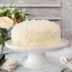 A coconut cake covered in shredded coconut sits on a white cake stand, with flowers and a teacup set in the background.