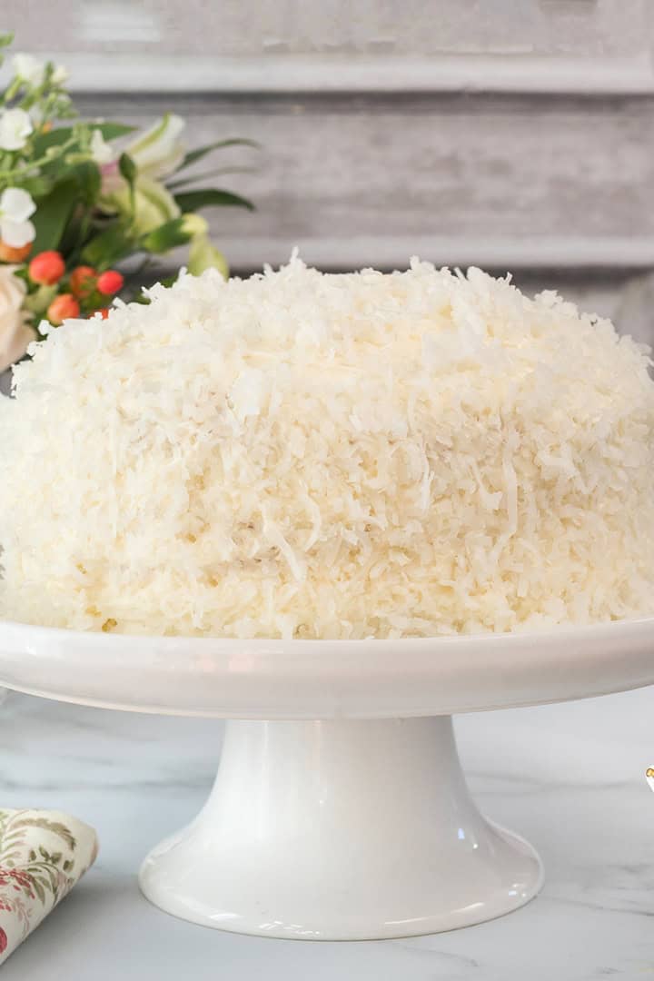 A coconut cake covered in shredded coconut sits on a white cake stand, with flowers and a teacup set in the background.