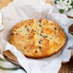 Round loaf of golden-brown spotted dog Irish soda bread with visible raisins, placed on parchment paper in a wicker basket.