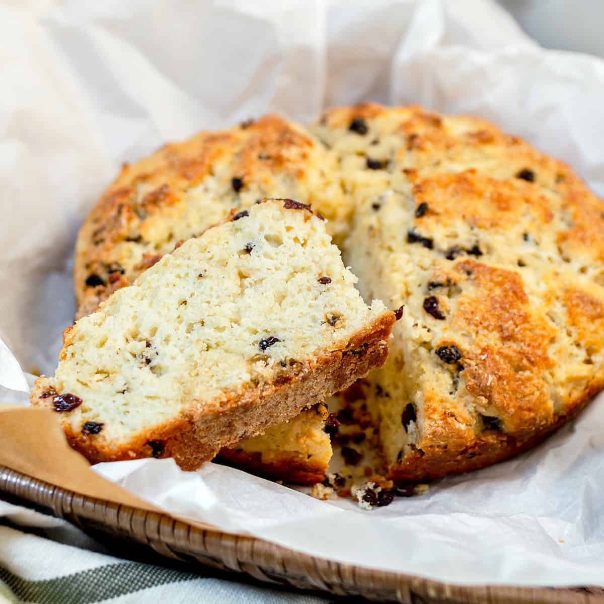 A round loaf of bread with raisins is partially sliced, with one wedge lifted to show the texture. The bread sits on parchment paper in a basket, ready to enjoy as the season begins.