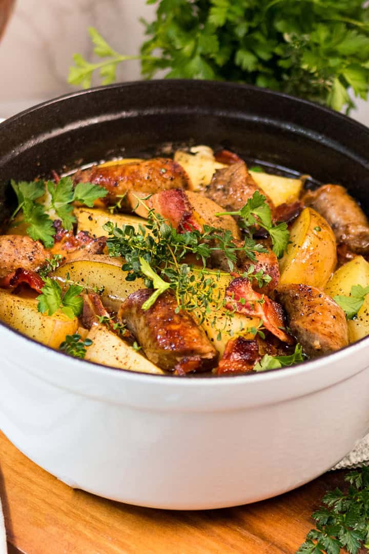 A white Dutch oven filled with Dublin Coddle-cooked chicken, potatoes, bacon, and fresh herbs-sits on a wooden surface with parsley nearby.