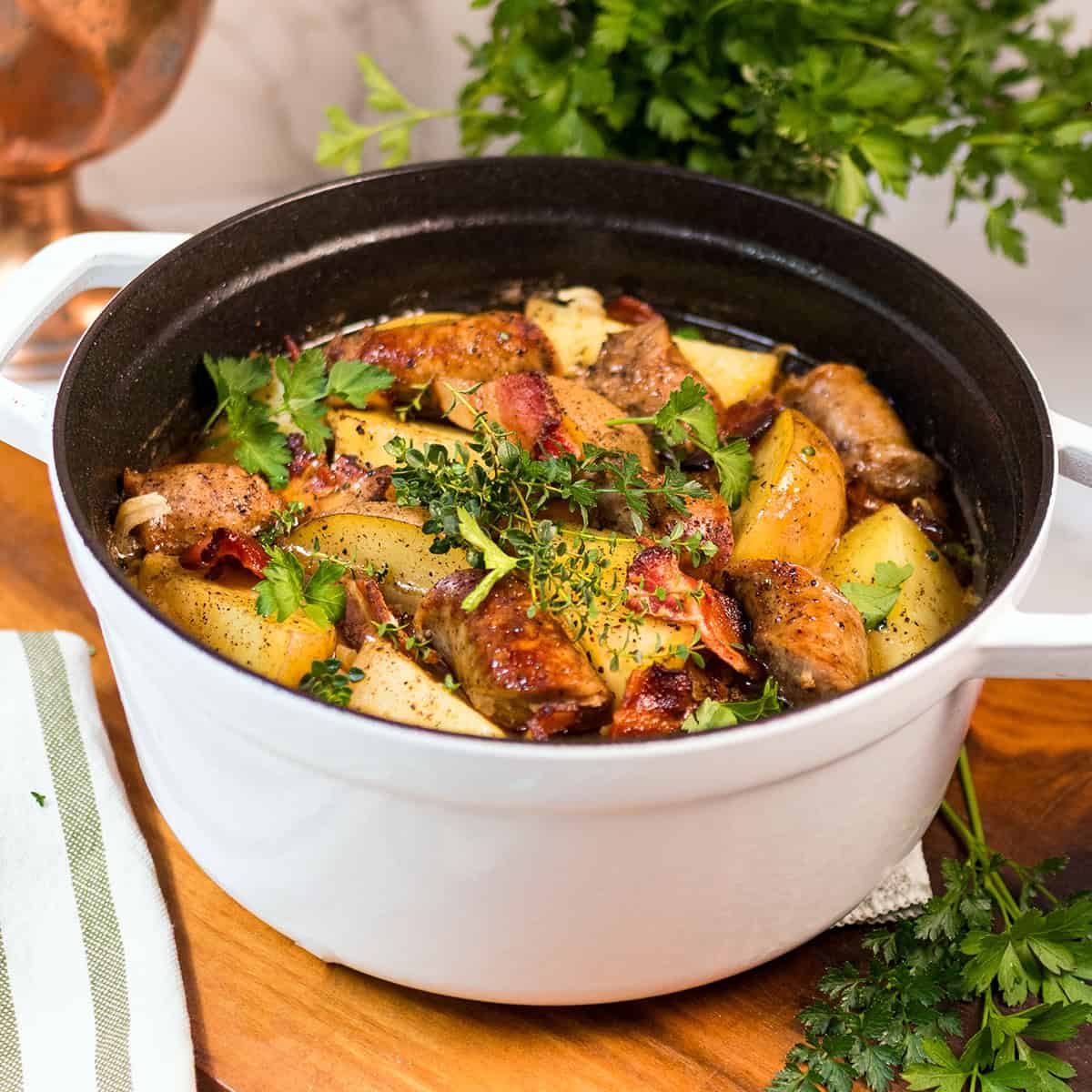 A white Dutch oven filled with Dublin Coddle-cooked chicken, potatoes, bacon, and fresh herbs-sits on a wooden surface with parsley nearby.