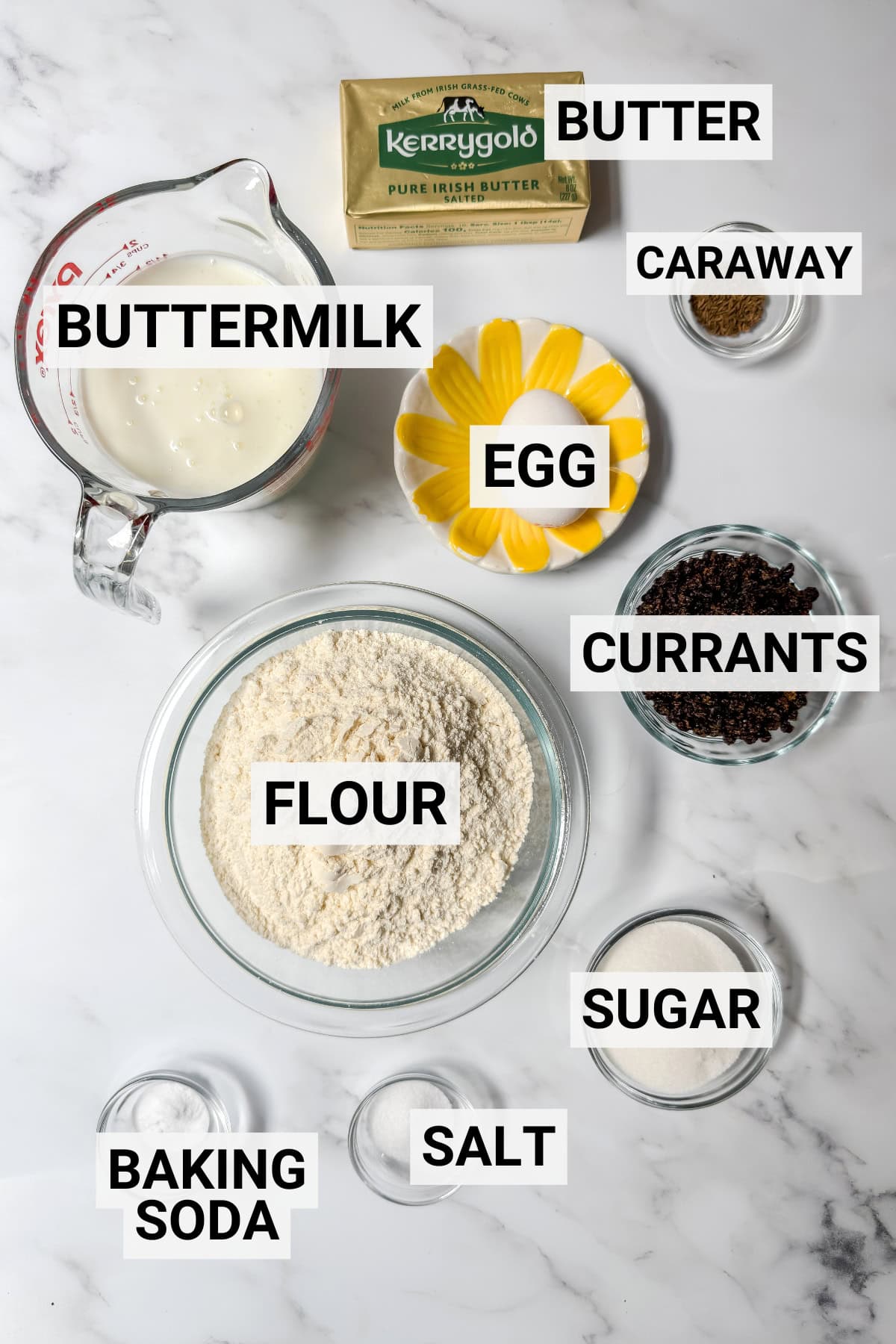 Baking ingredients for spotted dog Irish soda bread on a marble surface, including buttermilk, butter, caraway, egg, currants, flour, sugar, baking soda, and salt-each item neatly labeled.