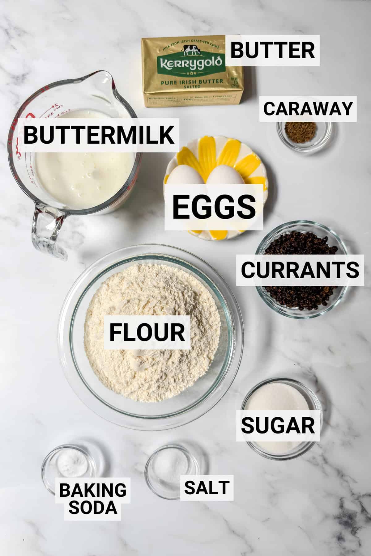 Ingredients for spotted dog Irish soda bread are arranged on a countertop, including butter, buttermilk, eggs, caraway, currants, flour, sugar, baking soda, and salt in labeled containers.