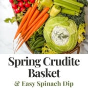 A basket filled with fresh spring vegetables, including carrots, celery, cauliflower, radishes, kale, and cabbages, served with spinach dip in cabbage bowls. Text reads "Spring Crudités Basket & Easy Spinach Dip.