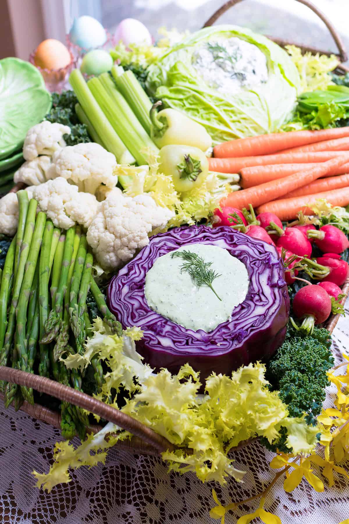 A basket filled with fresh spring vegetables including carrots, celery, asparagus, cauliflower, radishes, cabbage, and a purple cabbage half holding a creamy dip&mdash;perfect for serving as rustic crudit&eacute;s on a lace tablecloth.