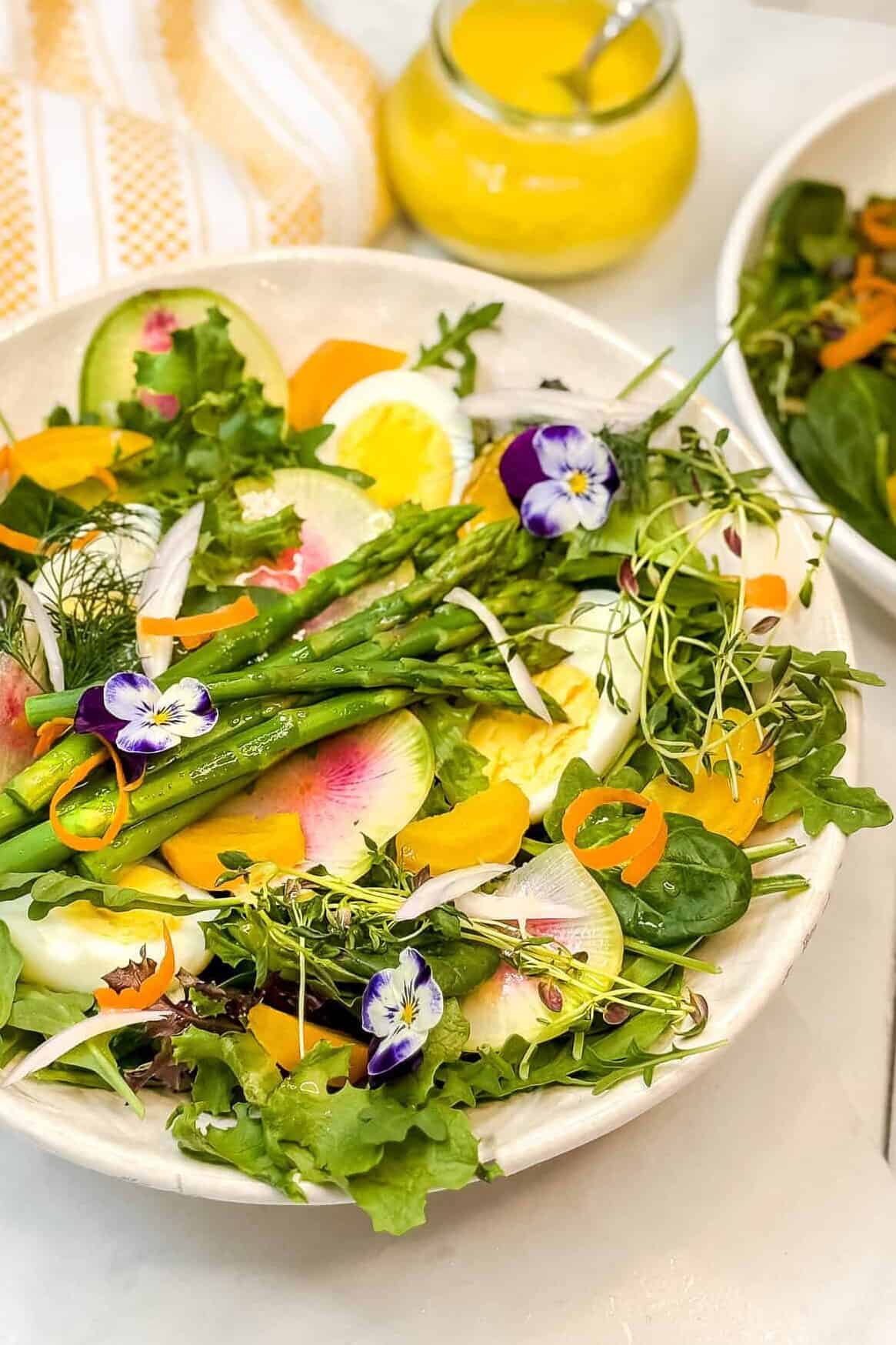 A vibrant salad with mixed greens, asparagus, sliced radishes, hard-boiled egg slices, and edible flowers. A small jar of yellow dressing with a spoon sits in the background on a white table with a striped cloth.