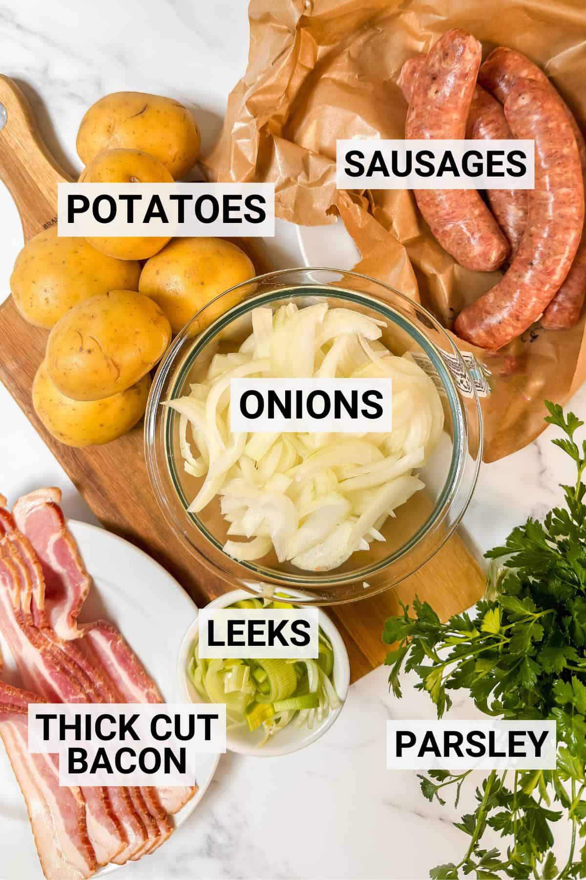 Sliced onions, potatoes, sausages, thick cut bacon, leeks, and parsley-classic ingredients for Dublin Coddle-are arranged on a white surface, each labeled with bold text.