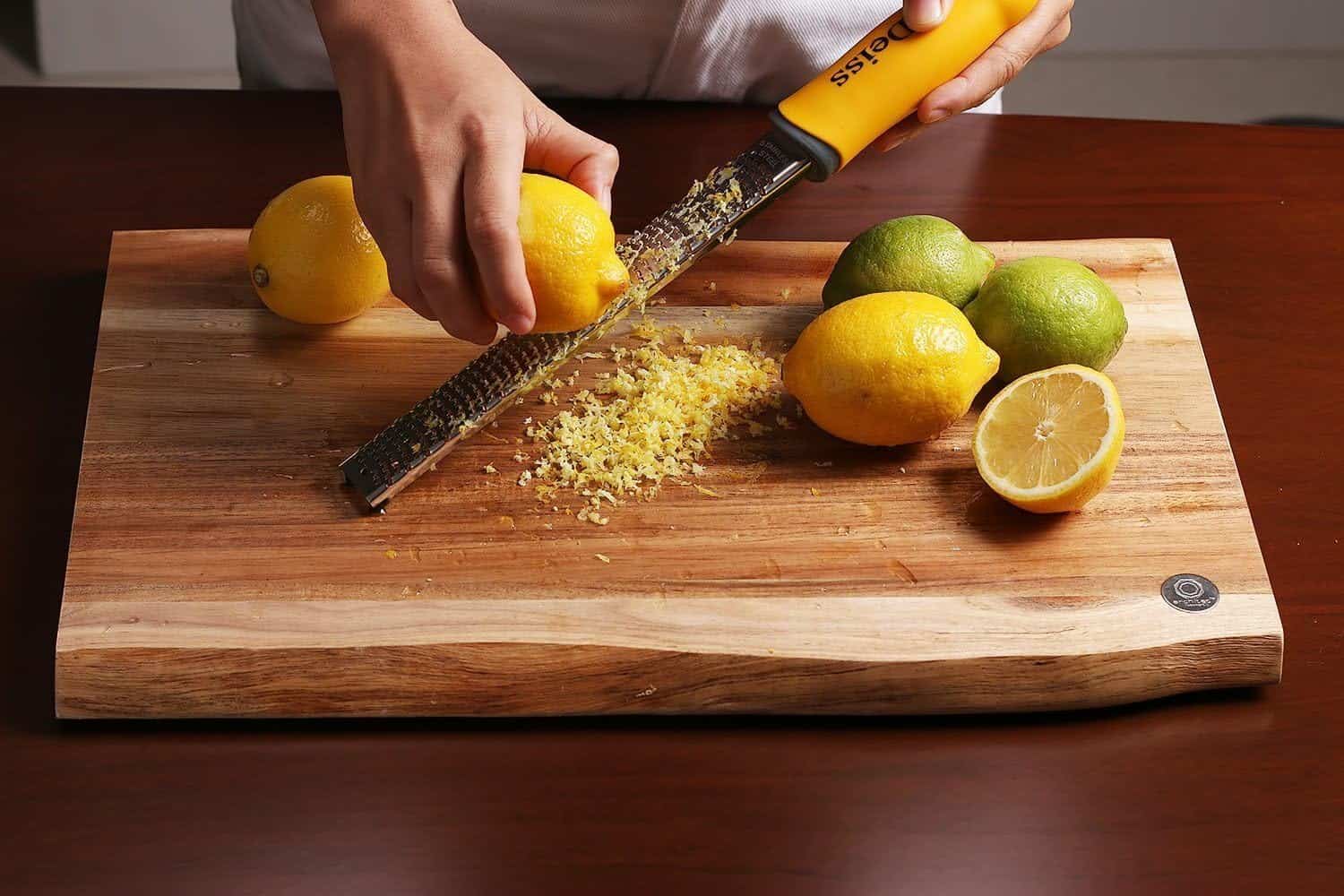 A person zesting a lemon with a grater on a wooden cutting board, surrounded by whole lemons and limes as the season begins.