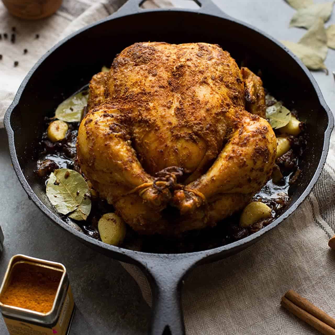 A whole roasted chicken seasoned with spices, placed in a black cast iron skillet with garlic cloves and bay leaves-because the season begins with comforting, flavorful meals.