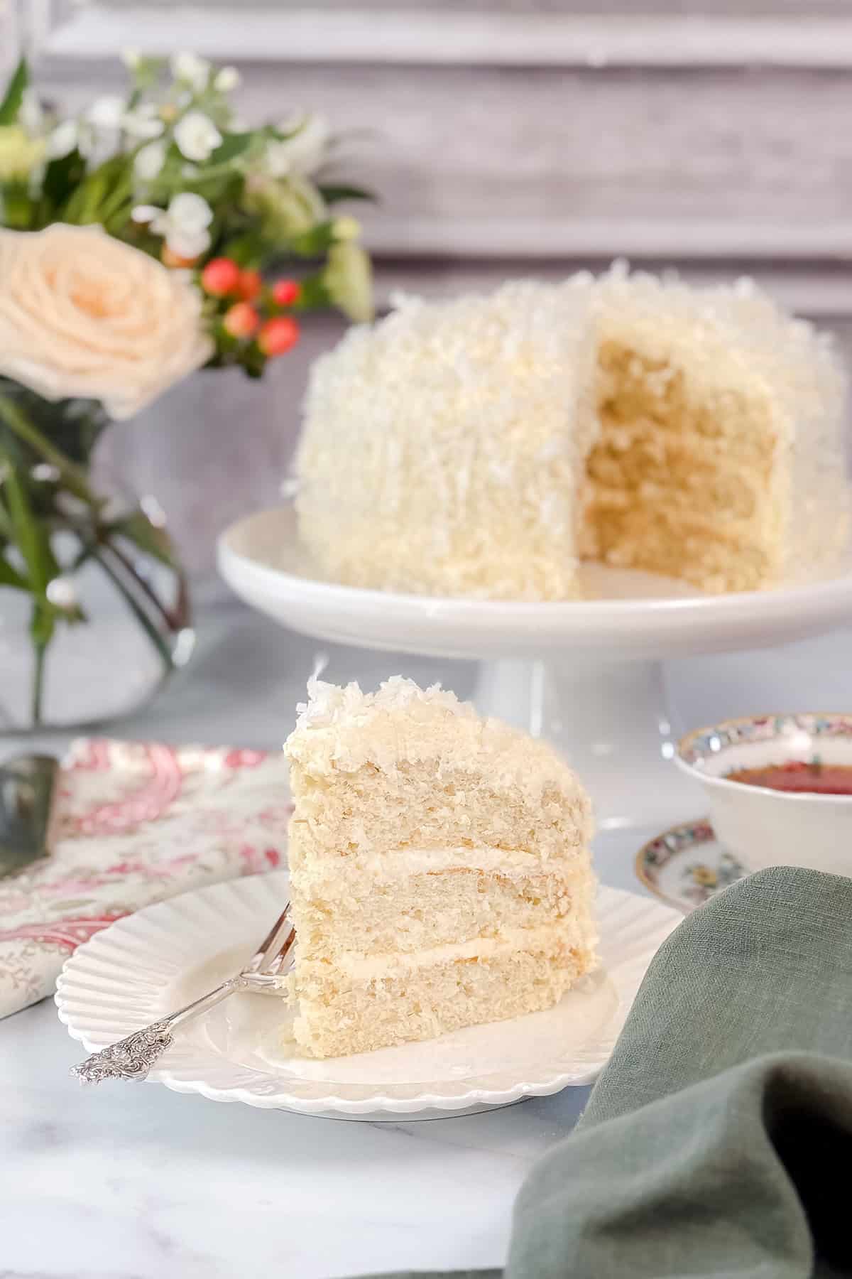 A slice of white frosted coconut cake sits on a plate with a fork; the rest of the cake is on a stand in the background next to a floral arrangement and tea cup.