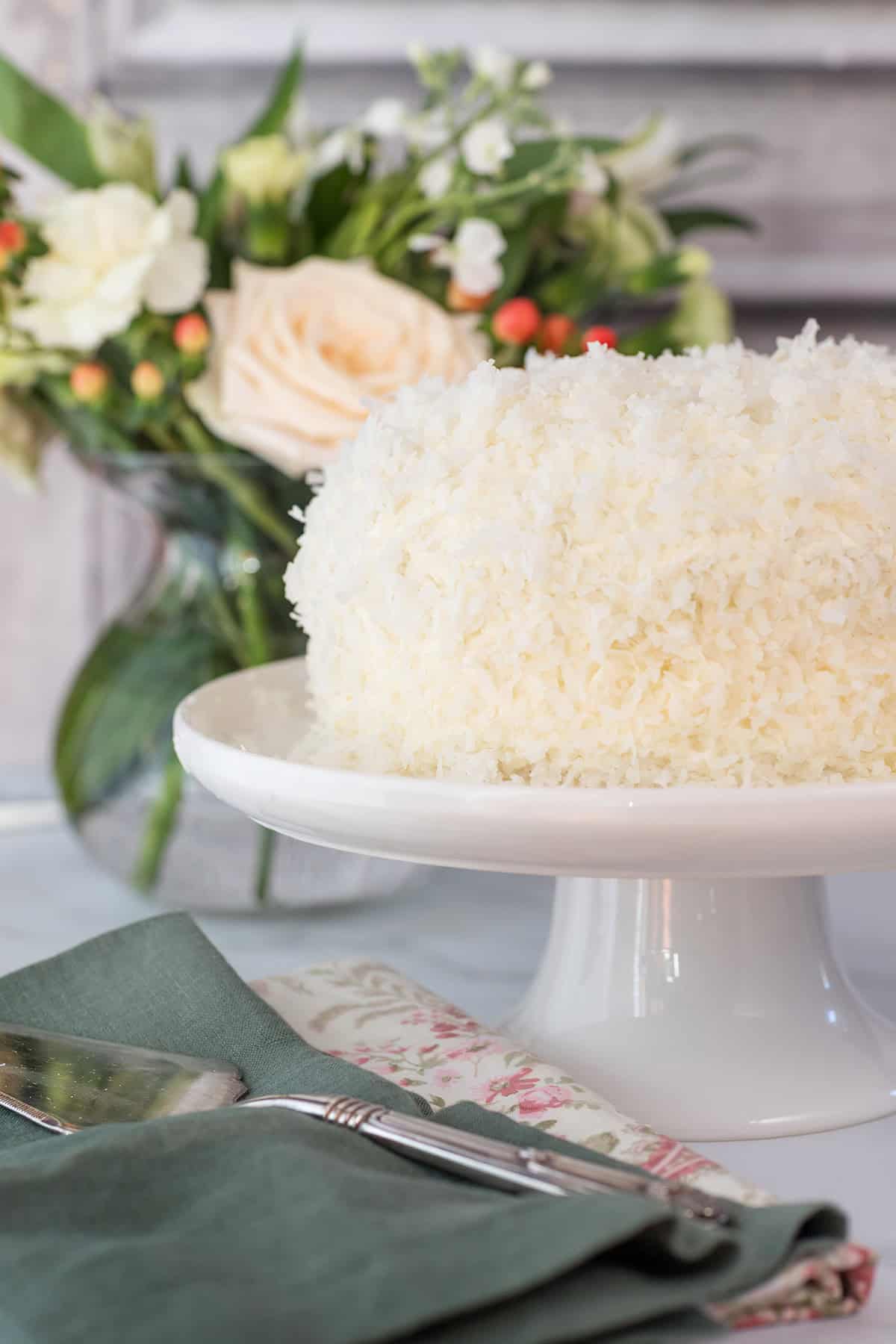 A white frosted coconut cake covered in shredded coconut sits on a white cake stand, with a floral arrangement and serving utensils nearby.