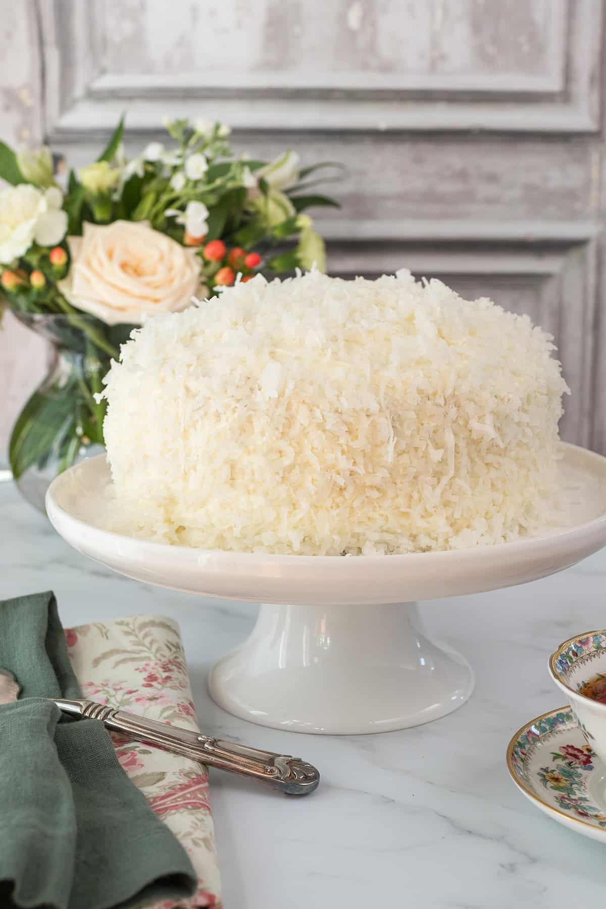 A round coconut cake covered in shredded coconut sits on a white cake stand, with a floral arrangement and plates in the background, making this coconut cake the perfect centerpiece for any celebration.
