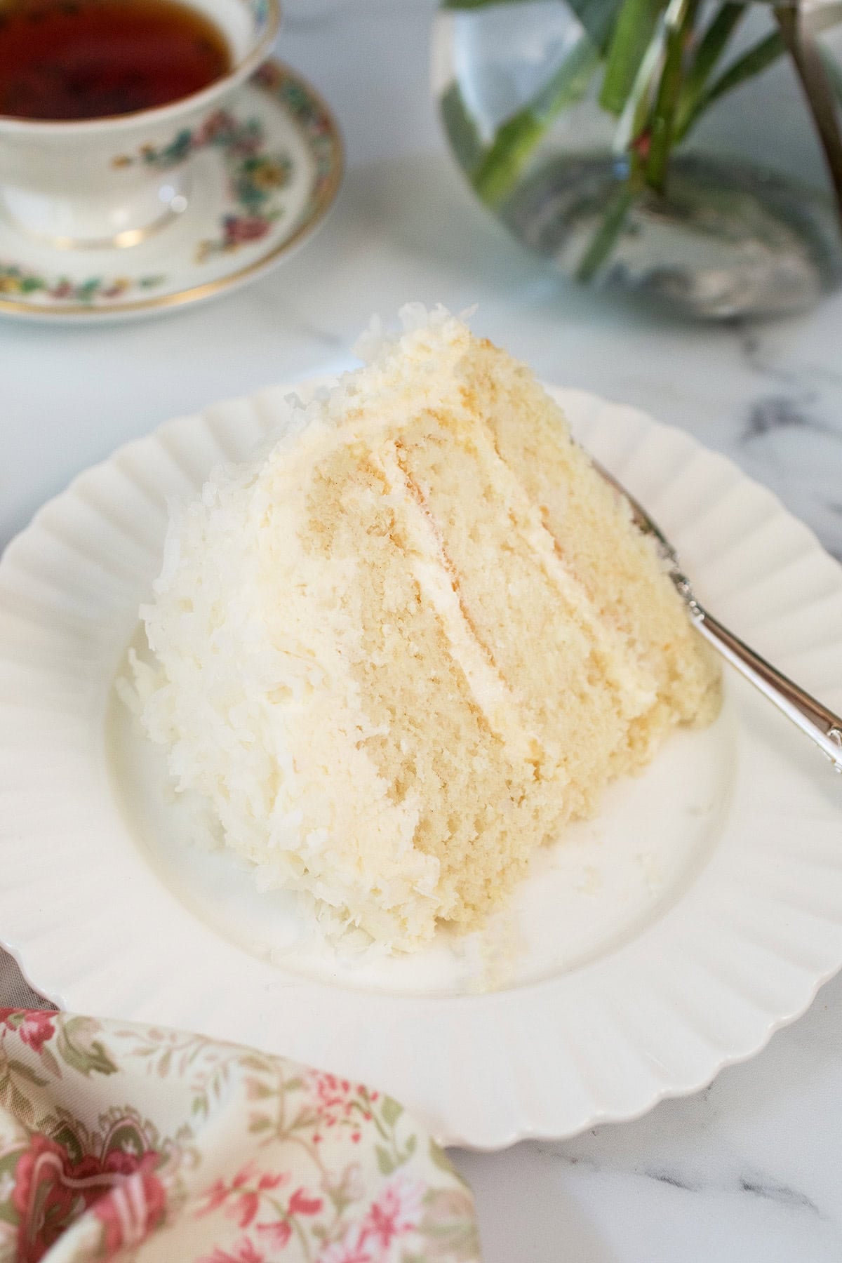 A slice of layered coconut cake with white frosting sits on a white plate, accompanied by a fork; in the background, a cup of tea and a flower vase enhance the inviting scene.