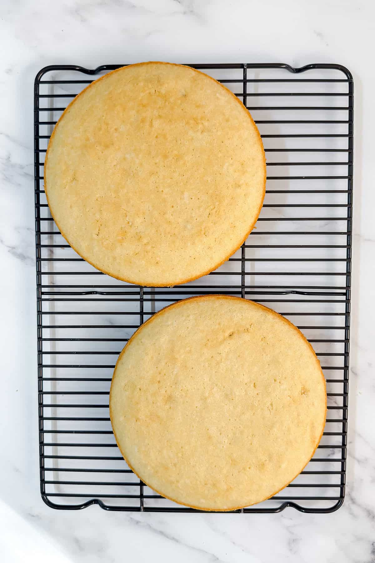 Two round coconut cakes cooling on a black wire rack placed on a white marble surface.