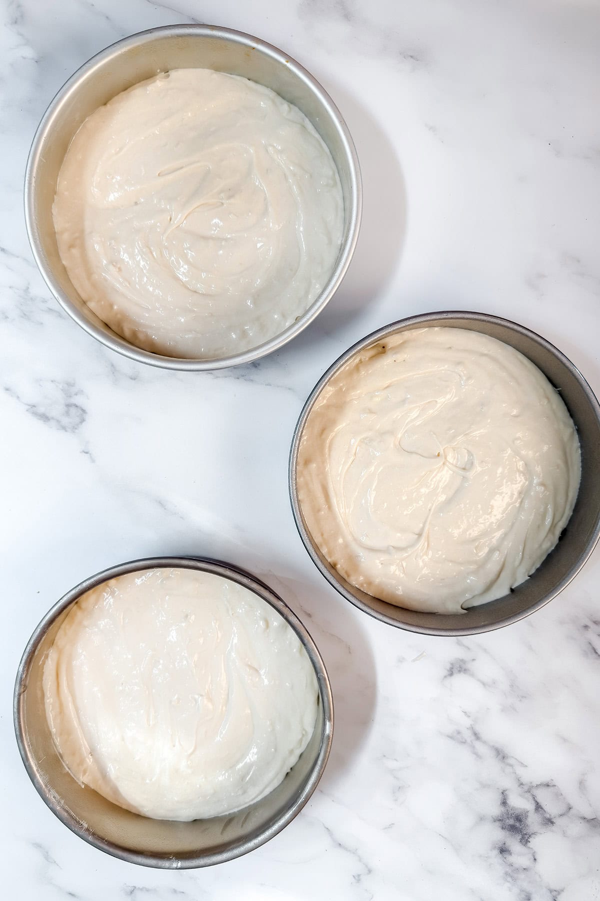 Three round cake pans filled with unbaked coconut cake batter are arranged on a white marble surface.