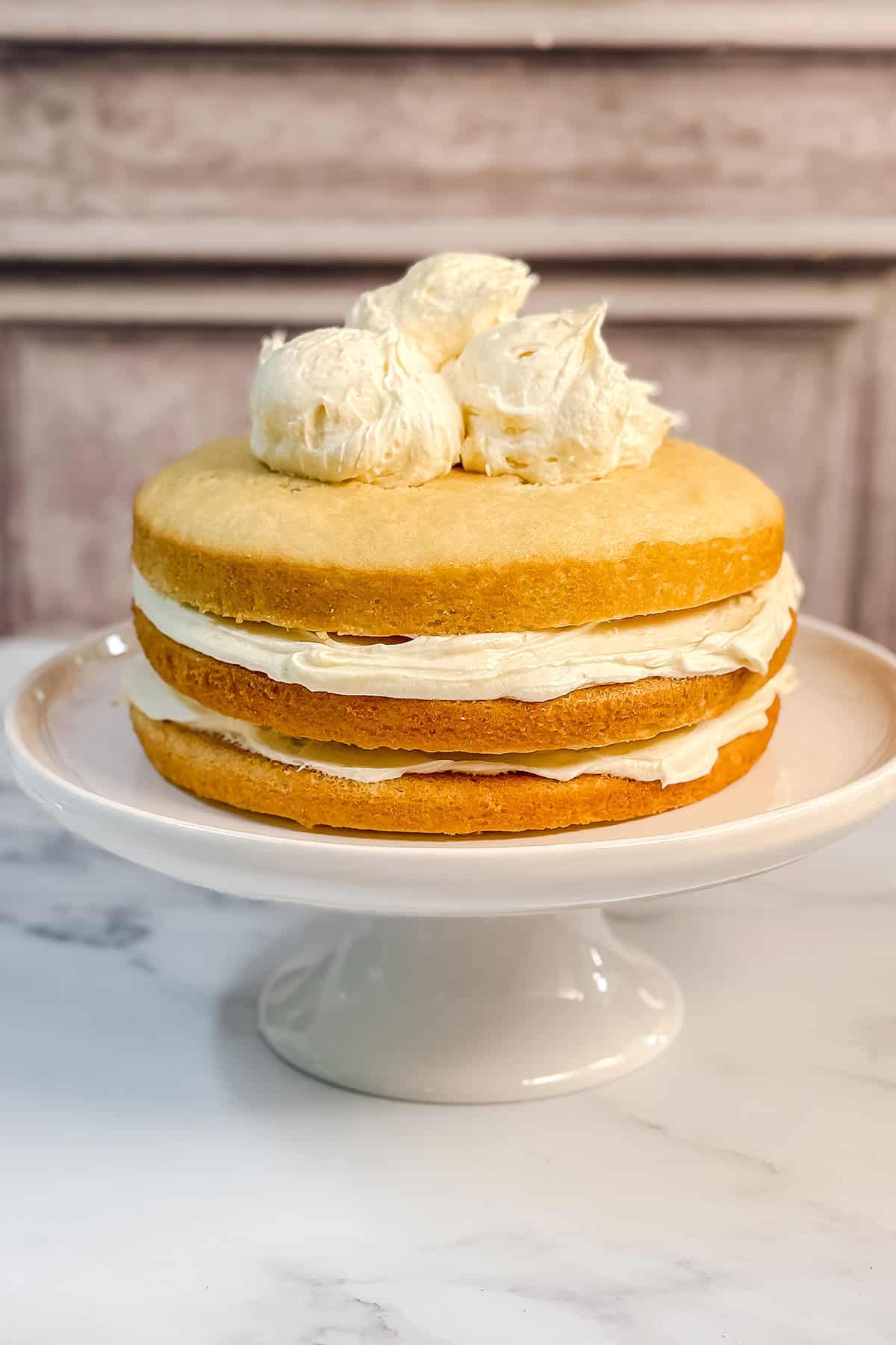 A three-layer coconut cake with with dollops of coconut cream cheese buttercream frosting on top to cover the cake entirely before sprinkling flaked coconut over the exterior.