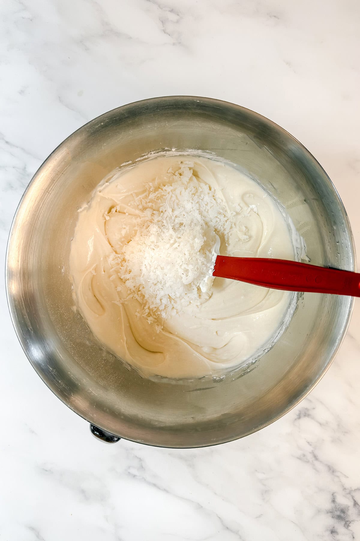 A metal mixing bowl containing creamy coconut cake batter with shredded coconut on top.