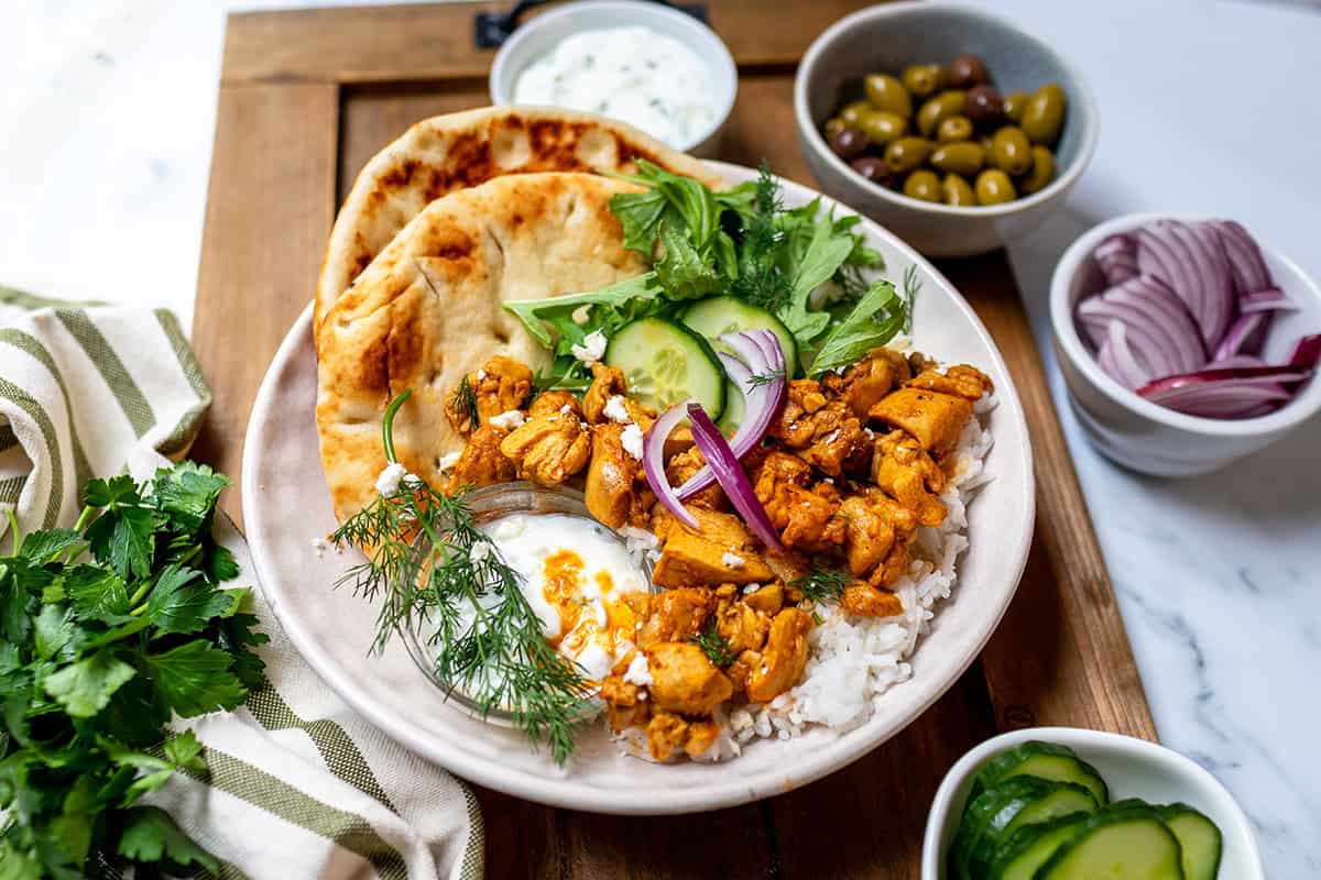 A plate with spiced chicken shawarma, rice, naan bread, cucumber, red onion, fresh herbs, yogurt sauce, and sides of olives and red onion slices on a wooden board-perfect for those who love quick recipes and flavorful shawarma bowls.