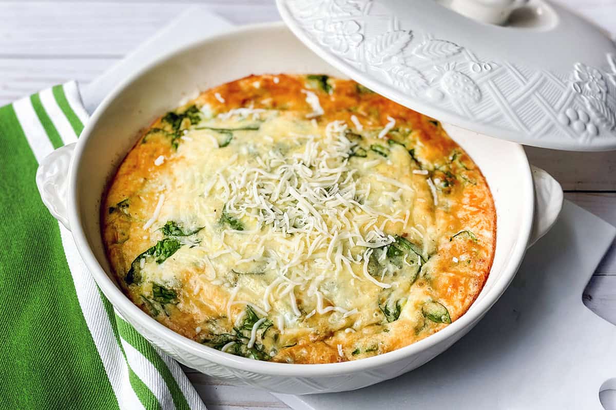 A baked frittata with spinach and melted cheese is served in a white casserole dish, partially covered, next to a green and white striped towel-perfect for fans of quick and easy recipes.