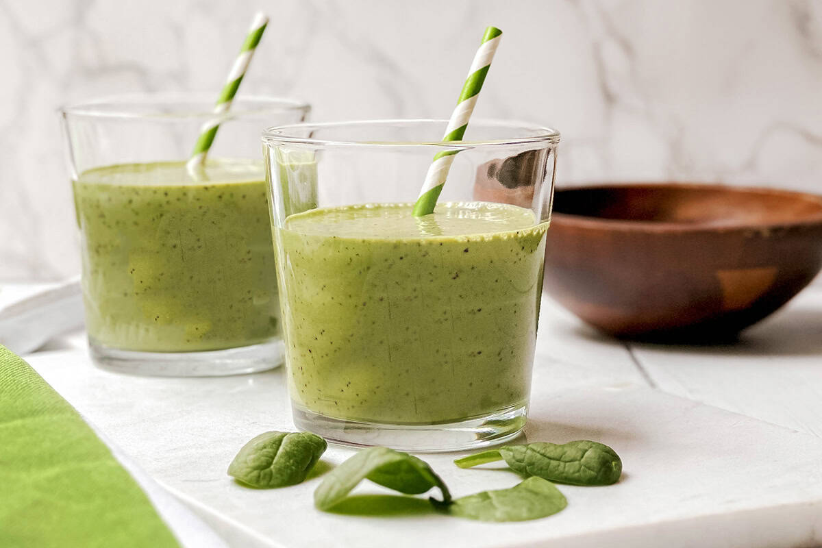 Two glasses of green smoothie with striped straws rest on a white surface, featuring fresh spinach leaves in front-perfect for enjoying your first greens-with a wooden bowl in the background.