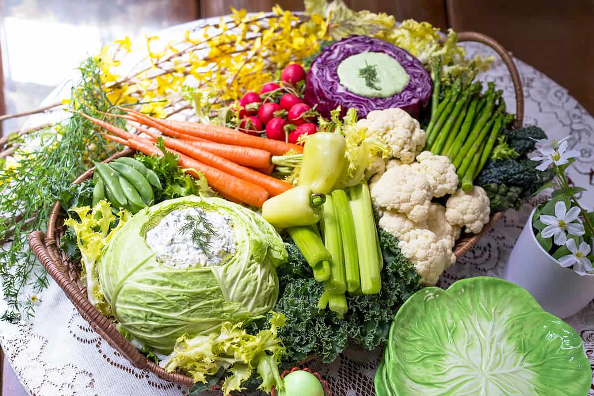 A basket on a lace tablecloth filled with fresh spring vegetables-cabbage, carrots, radishes, cauliflower, celery, and asparagus-creates a vibrant spring crudit&eacute;s display, surrounded by yellow flowers and a green plate.