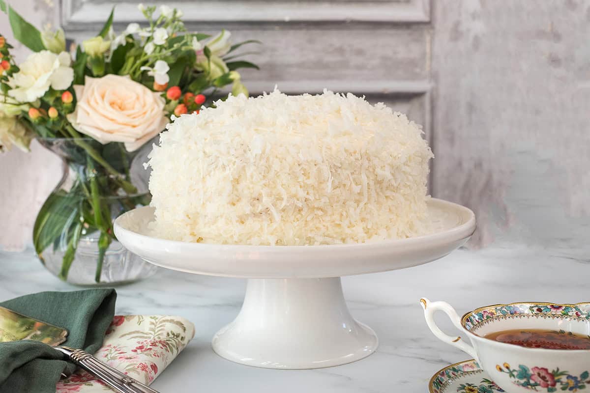 A layered coconut cake sits on a white cake stand, its surface covered in shredded coconut, with a floral arrangement and a cup of tea in the background.