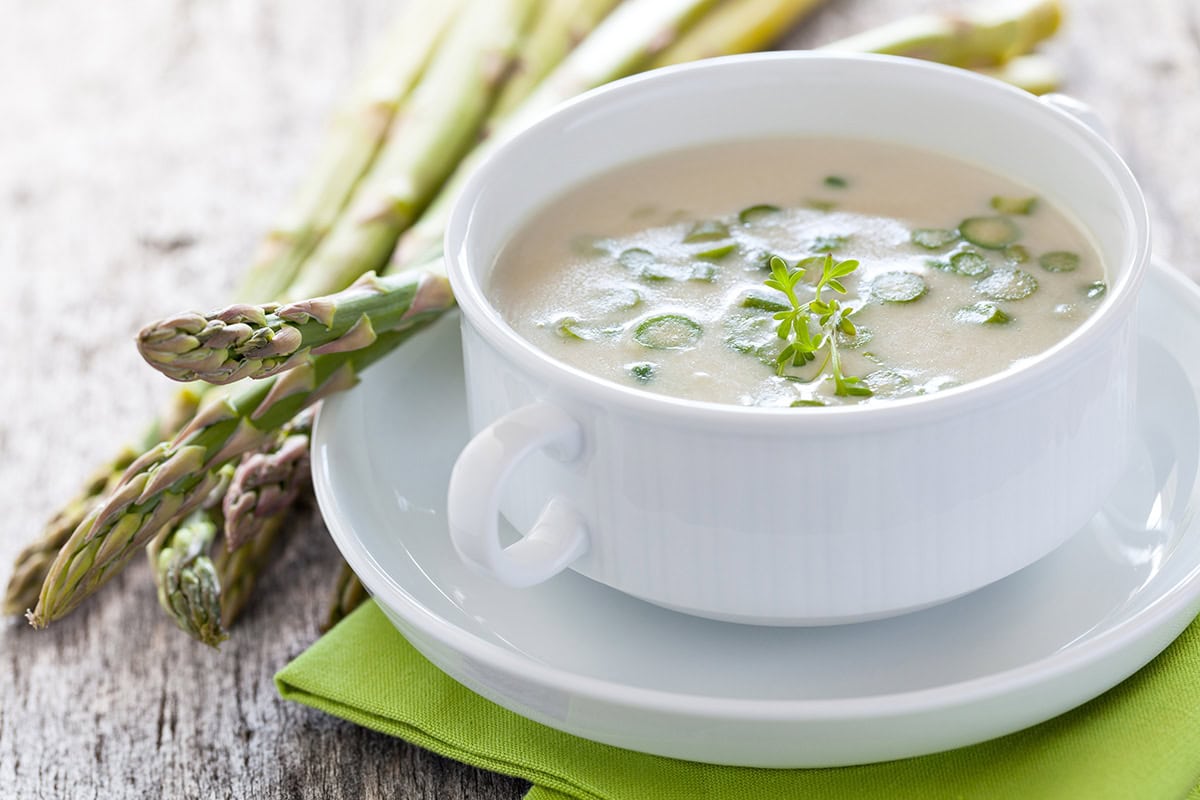 A white cup filled with creamy asparagus soup garnished with herbs sits on a matching saucer, next to fresh asparagus spears on a green napkin.