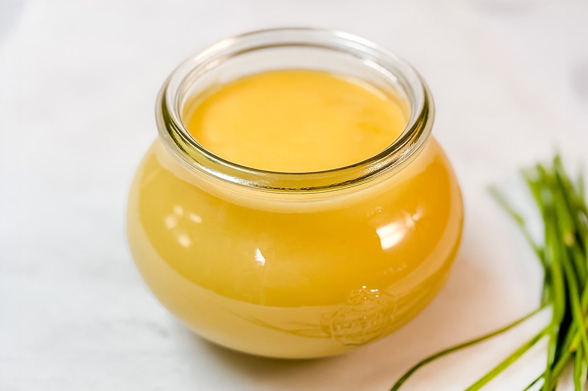 A glass jar filled with light yellow ghee sits on a white surface, with some green chives visible on the right side-perfect for crafting stovetop hollandaise sauce or as a rich base for your favorite sauce.