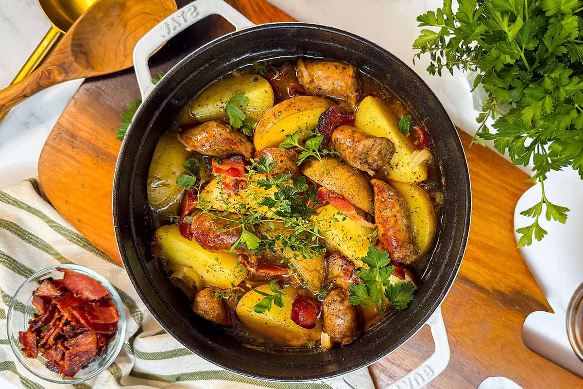 A pot of Dublin Coddle filled with roasted potatoes, sausage, tomatoes, and herbs, garnished with fresh parsley, sits on a wooden board next to a bowl of bacon and a bunch of parsley.