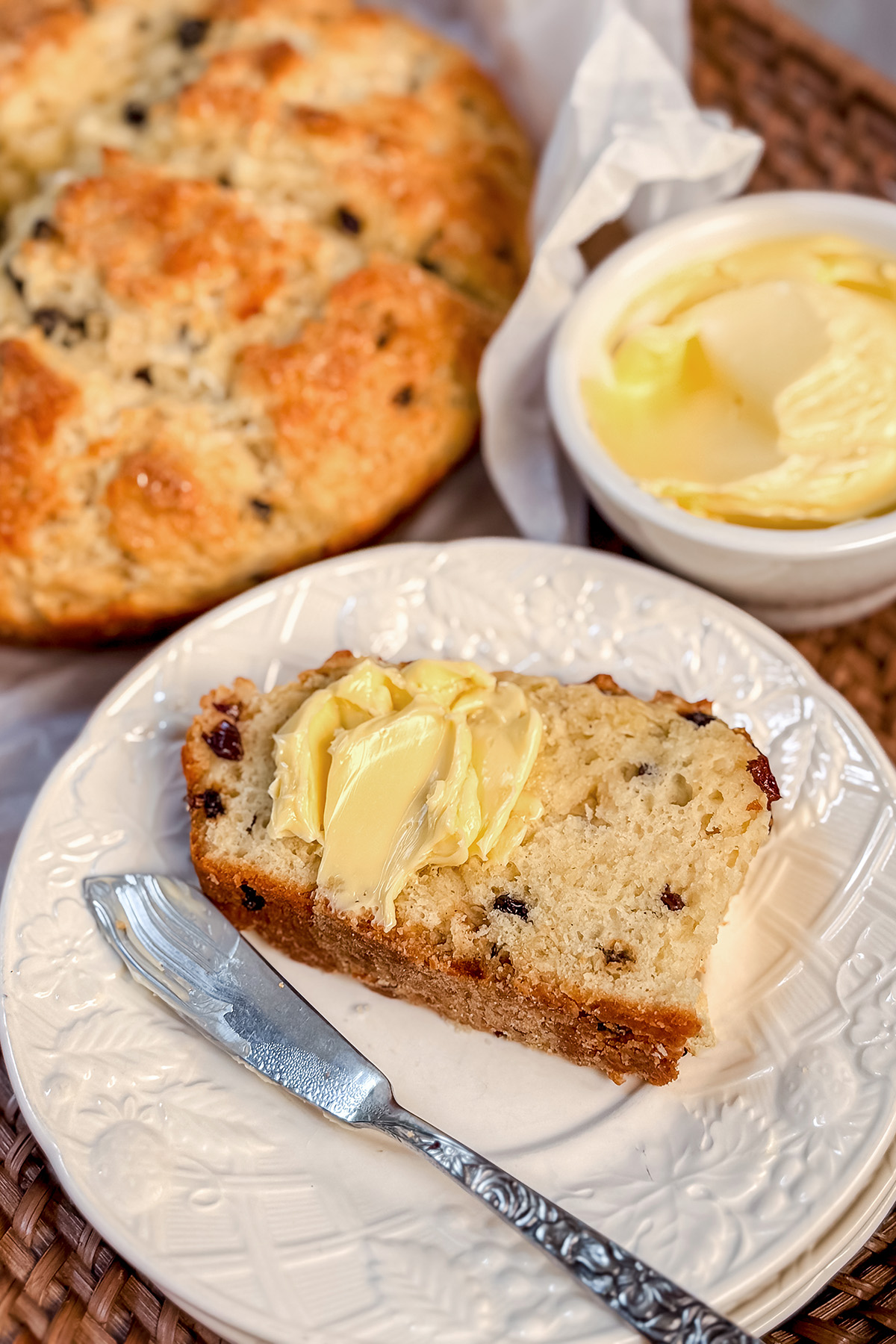 A slice of spotted dog Irish soda bread with a pat of butter on a white plate, a knife alongside, with a butter dish and more bread in the background.