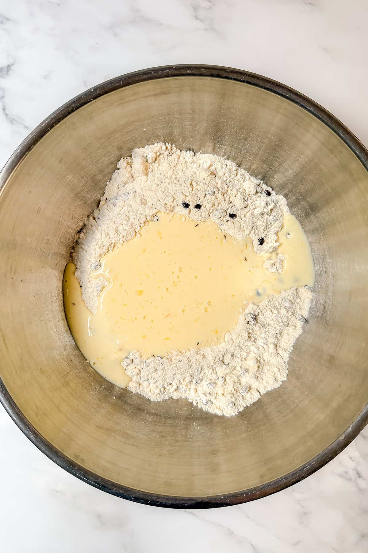 A large metal mixing bowl containing flour mixed with raisins, ready for spotted dog Irish soda bread, with a well in the center filled with liquid ingredients.