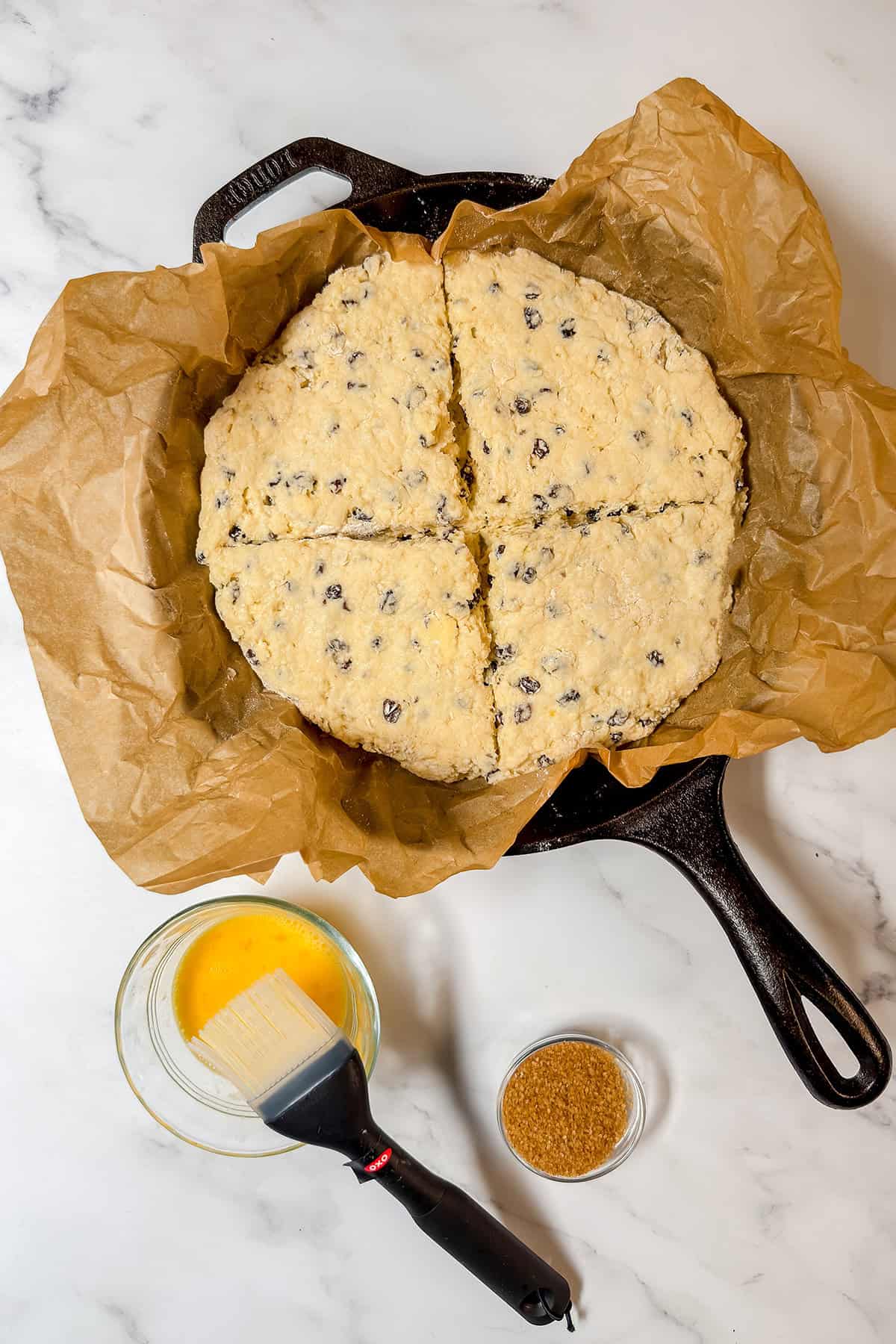 Unbaked spotted dog Irish soda bread dough with raisins sits in a parchment-lined cast iron skillet, scored into four pieces, with a bowl of egg wash, pastry brush, and small bowl of sugar nearby.