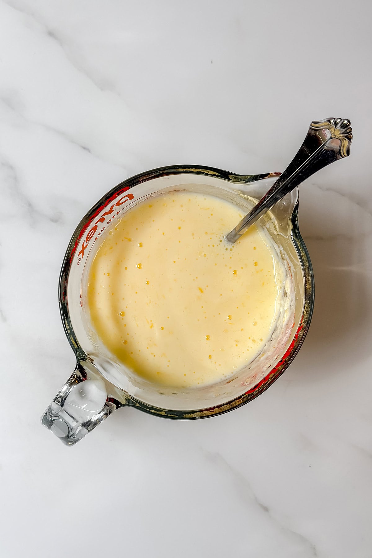 A glass measuring cup filled with a pale yellow mixture, likely for making spotted dog Irish soda bread, with a metal spoon inside, placed on a white marble surface.