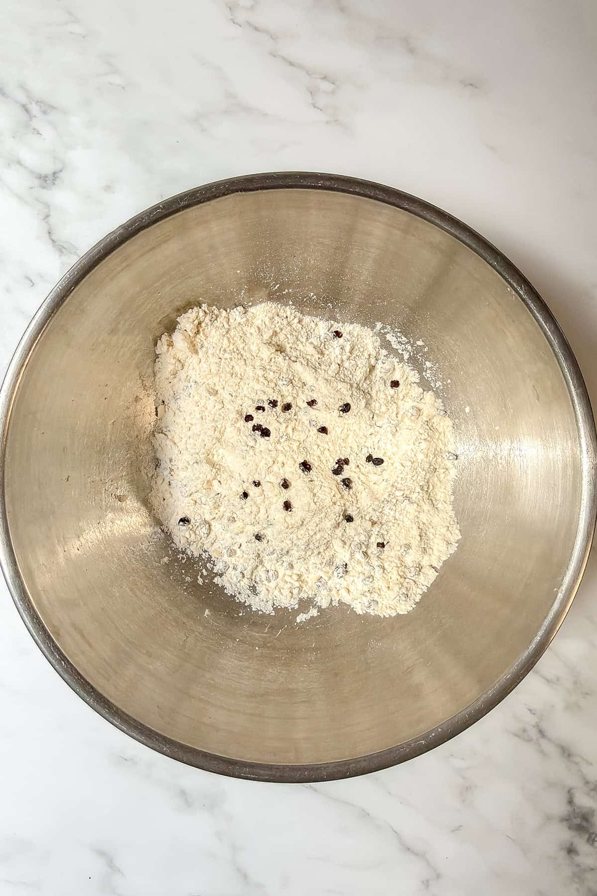 A metal mixing bowl containing flour mixture with small dark specks, likely dried fruit or seeds, sits on a white marble surface-perfect for making spotted dog Irish soda bread.