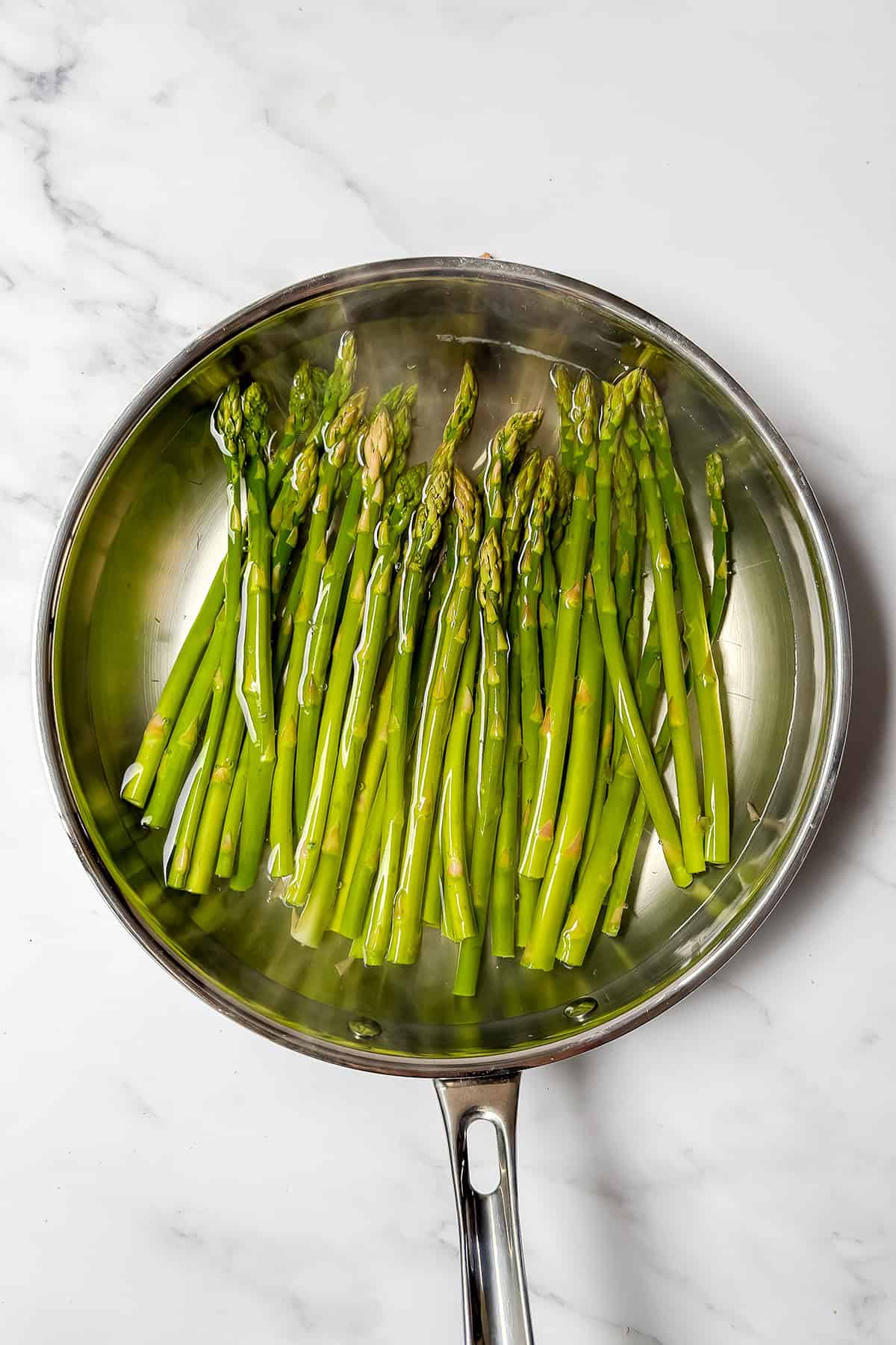 Fresh spring vegetables like asparagus spears are cooked in a stainless steel skillet with water on a white marble surface, perfect for adding to your Spring Crudites spread.