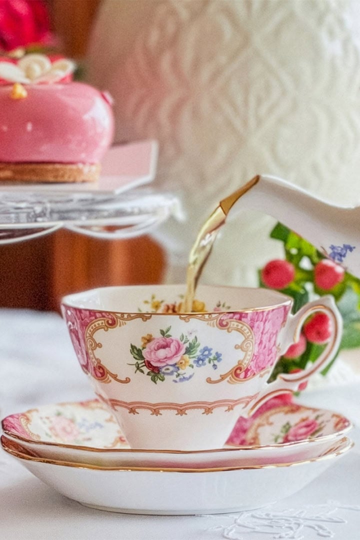 Tea is being poured from a teapot into a floral teacup, beside a pink dessert, strawberries, and a white patterned vase-perfect for a Regency afternoon tea inspired by Jane Austen.