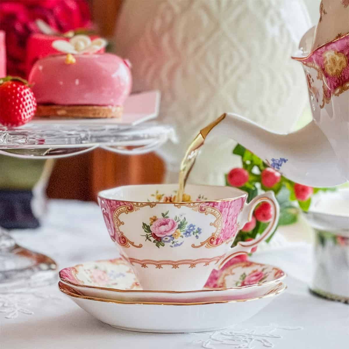 Tea is being poured from a teapot into a floral teacup, beside a pink dessert, strawberries, and a white patterned vase-perfect for a Regency afternoon tea inspired by Jane Austen.