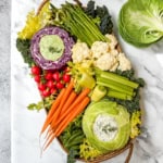 A vibrant Spring Crudités platter featuring fresh carrots, radishes, celery, asparagus, cauliflower, and lettuce, with dip elegantly served in hollowed cabbage and red cabbage halves.