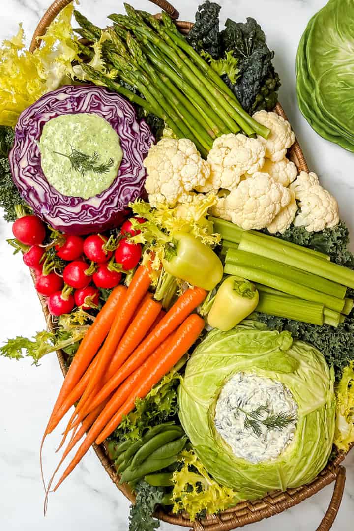 A vibrant Spring Crudités platter featuring fresh carrots, radishes, celery, asparagus, cauliflower, and lettuce, with dip elegantly served in hollowed cabbage and red cabbage halves.