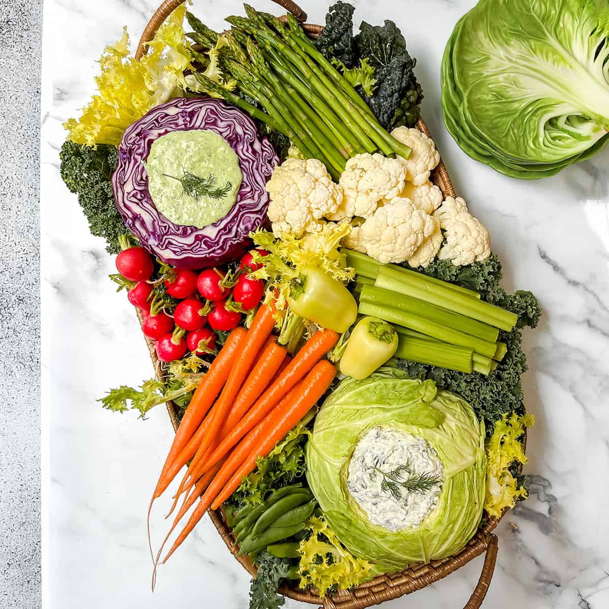 A vibrant Spring Crudités platter featuring fresh carrots, radishes, celery, asparagus, cauliflower, and lettuce, with dip elegantly served in hollowed cabbage and red cabbage halves.