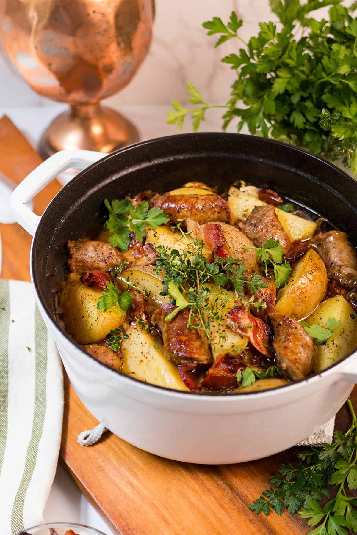 A white pot filled with Dublin Coddle&mdash;a stew of sausages, potatoes, herbs, and vegetables&mdash;sits on a wooden board, garnished with fresh parsley.