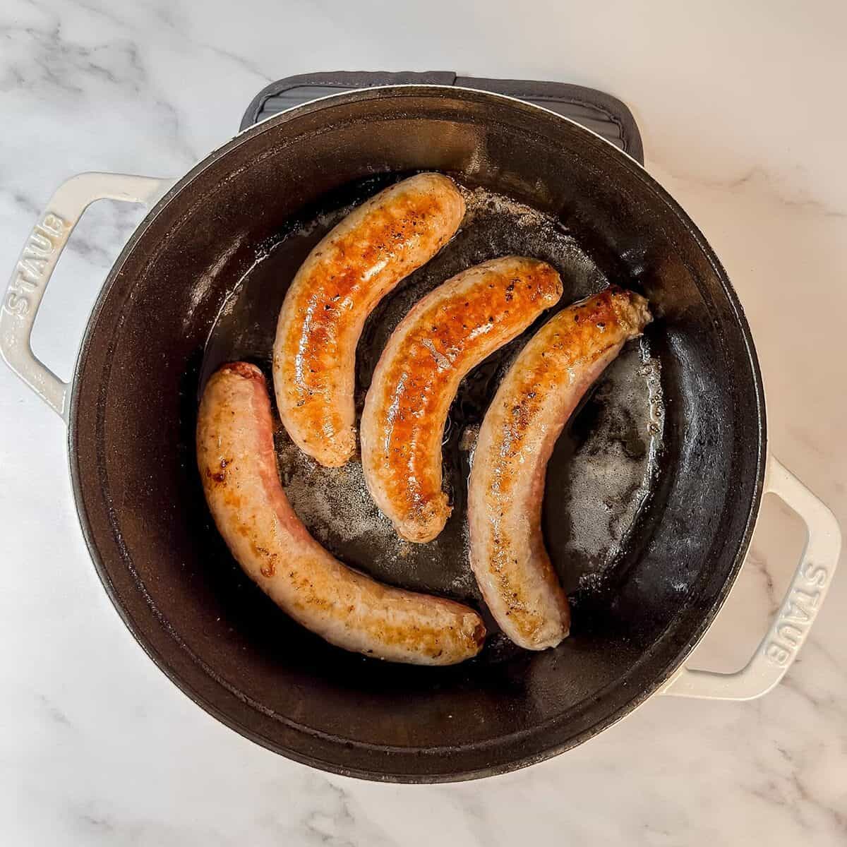 Four sausages are being cooked in a black cast iron pan on a white marble countertop, perfect for starting a hearty Dublin Coddle.