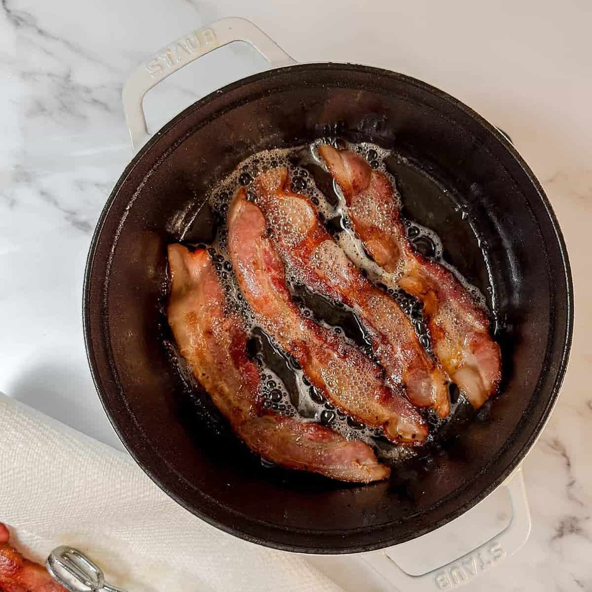 Strips of bacon frying in a black pot on a stovetop, perfect for starting a hearty Dublin Coddle, with cooked bacon resting on a paper towel and metal tongs beside it.