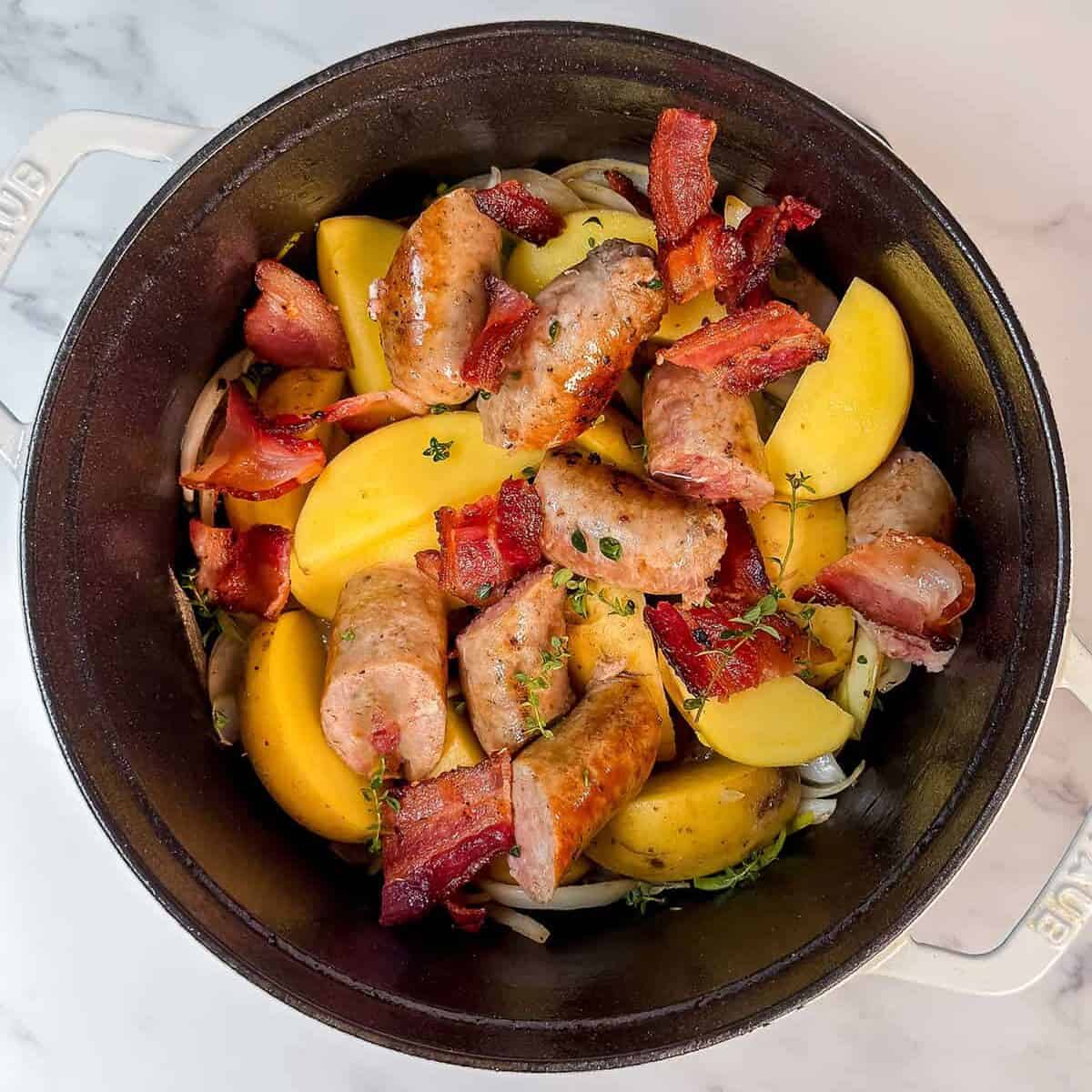 A Dutch oven filled with sausage links, bacon pieces, potato wedges, onion slices, and fresh herbs on a white marble surface-classic ingredients for a hearty Dublin Coddle.