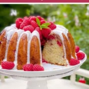 A full size White Chocolate Raspberry Bundt cake topped with white icing, fresh raspberries, and mint is displayed on a white cake stand outdoors—perfect among full size desserts for afternoon tea, with greenery in the background.