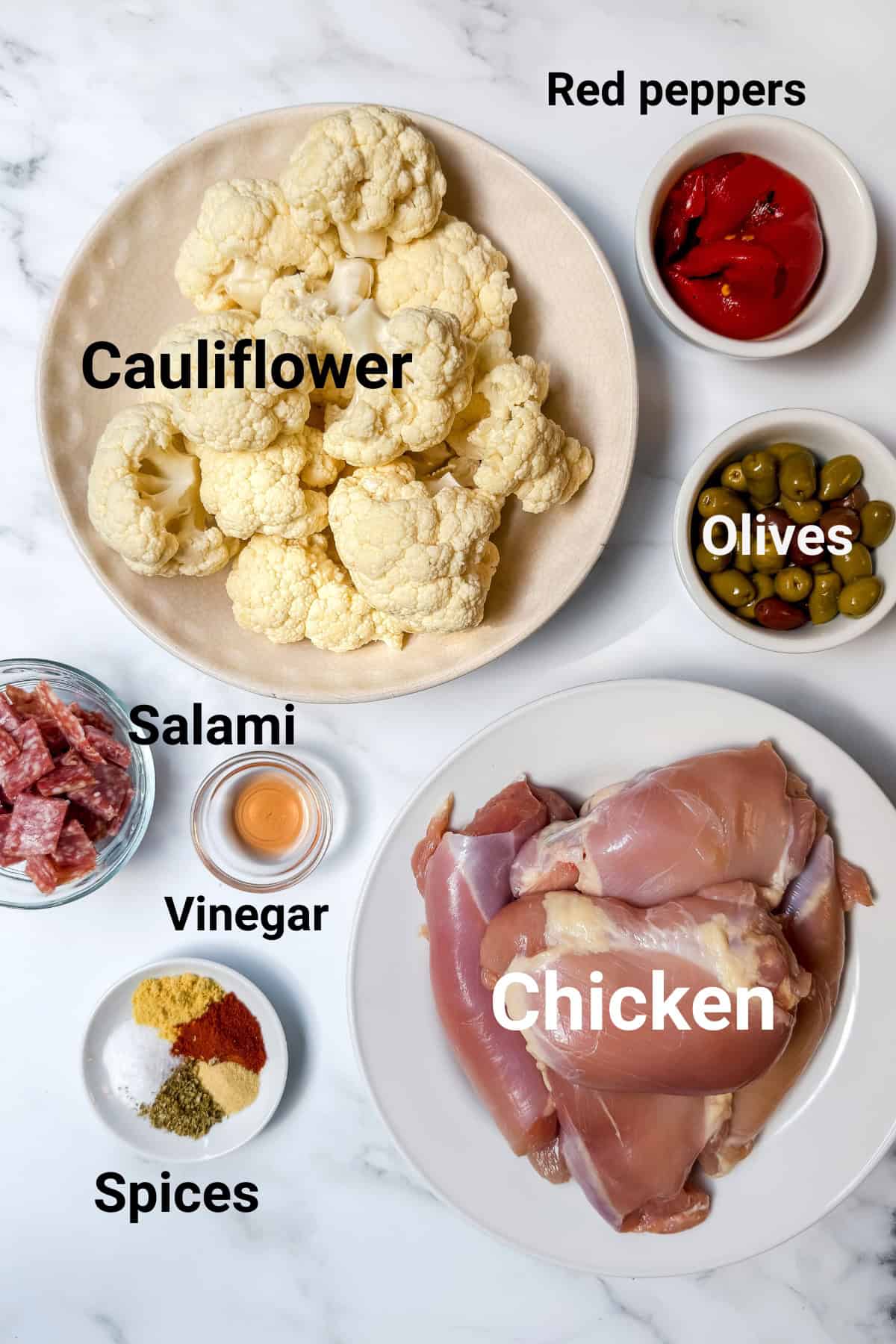 A flat lay of ingredients on a marble surface for a flavorful sheet pan chicken recipe: raw chicken thighs, cauliflower florets, red peppers, green olives, salami slices, vinegar, and assorted spices inspired by Muffaletta flavors.