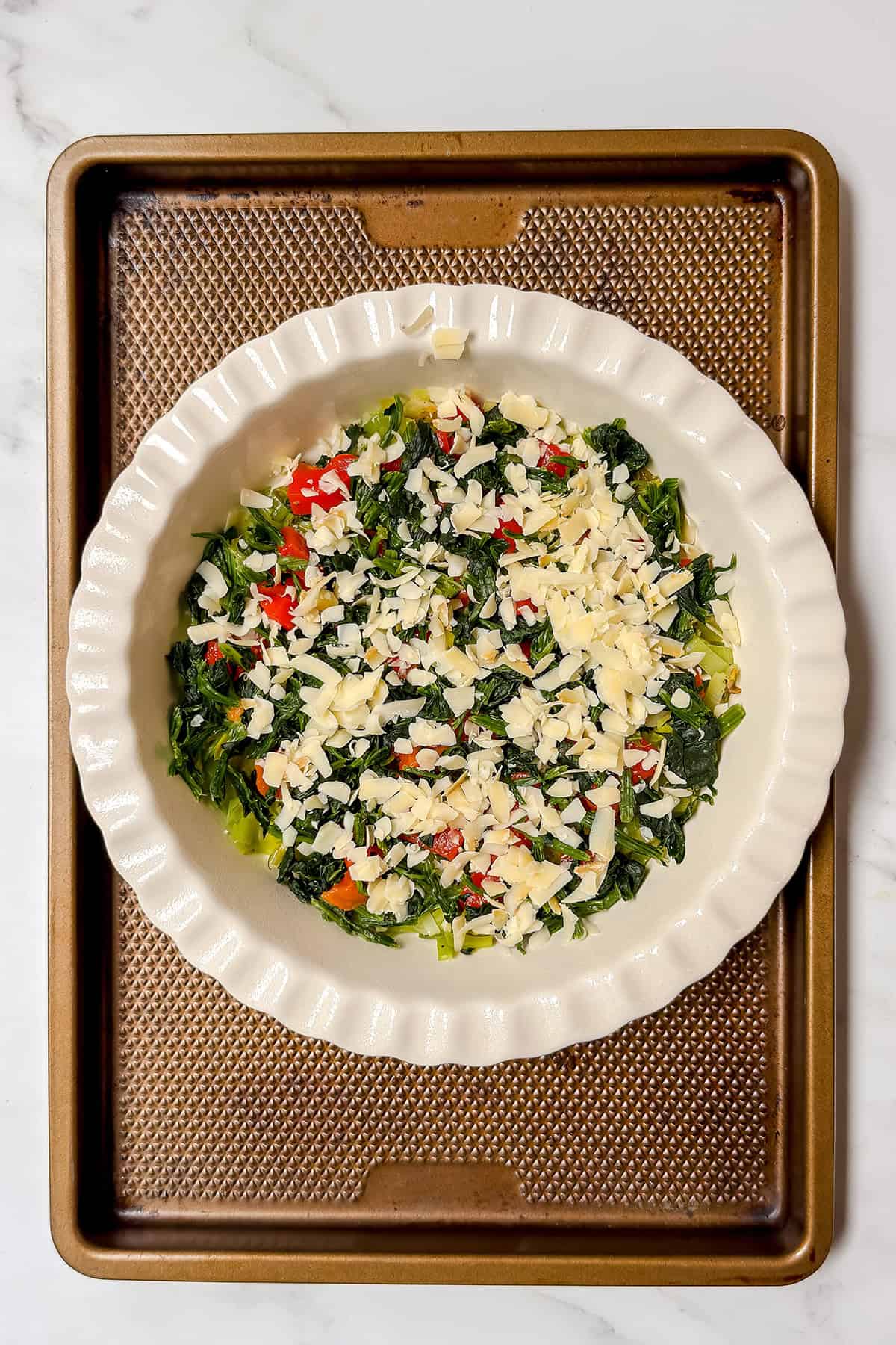 A white pie dish on a baking sheet holds a colorful crustless quiche filled with chopped spinach, red peppers, and shredded cheese on a marble countertop.