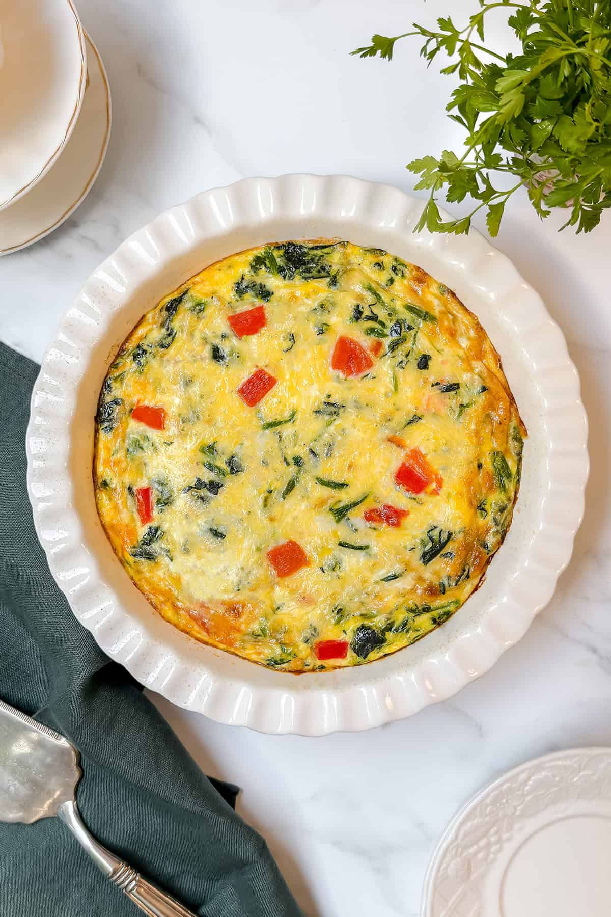 A crustless quiche packed with spinach and red peppers, baked in a white ceramic dish on a marble surface, sits beside a green napkin and fresh herbs.