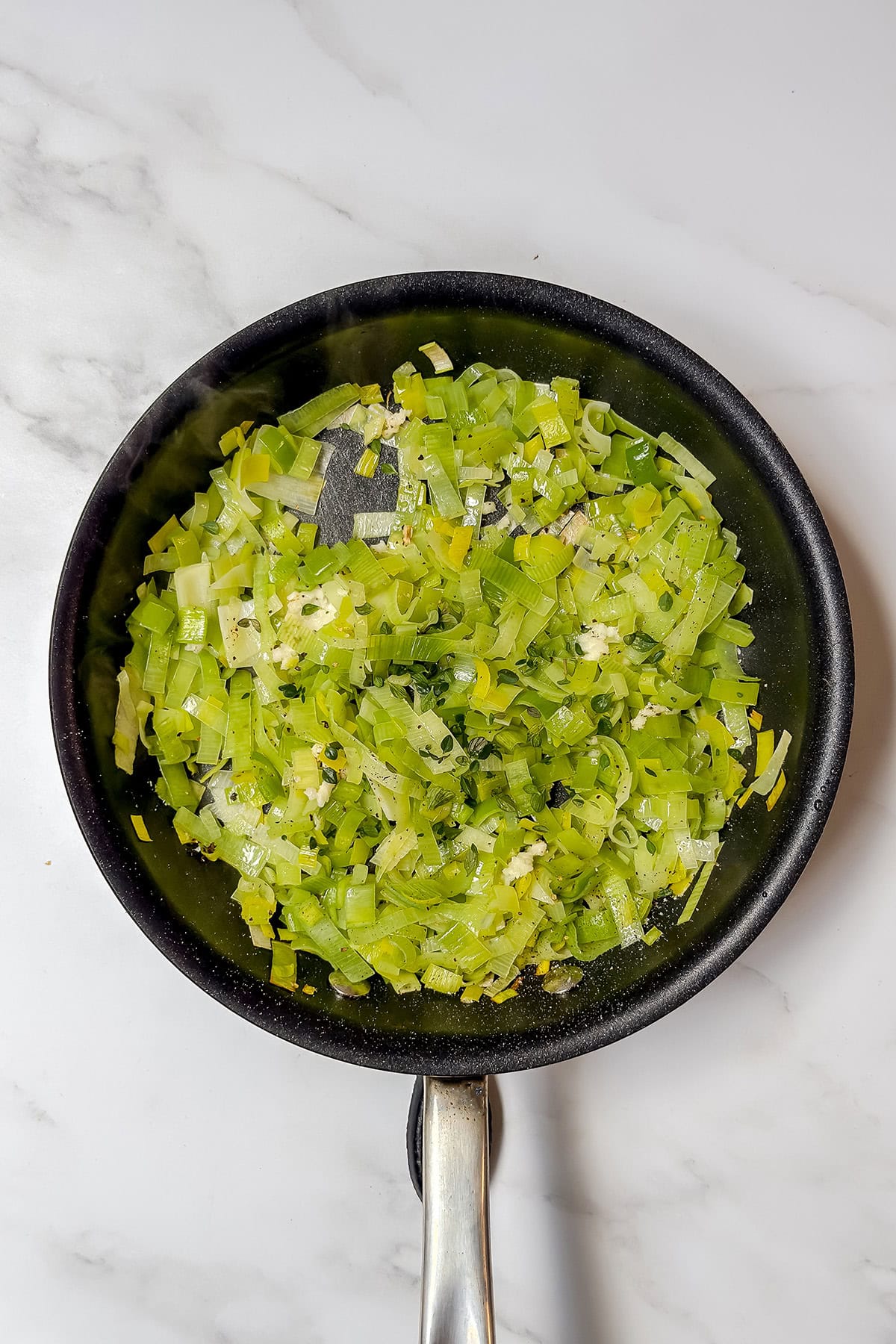 A black skillet containing saut&eacute;ed chopped leeks, minced garlic, and vibrant red peppers on a white marble surface.